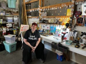 Woman sits in front of a workbench where she makes jewelry. Samples of the jewelry are hanging from a shelf above the workbench.